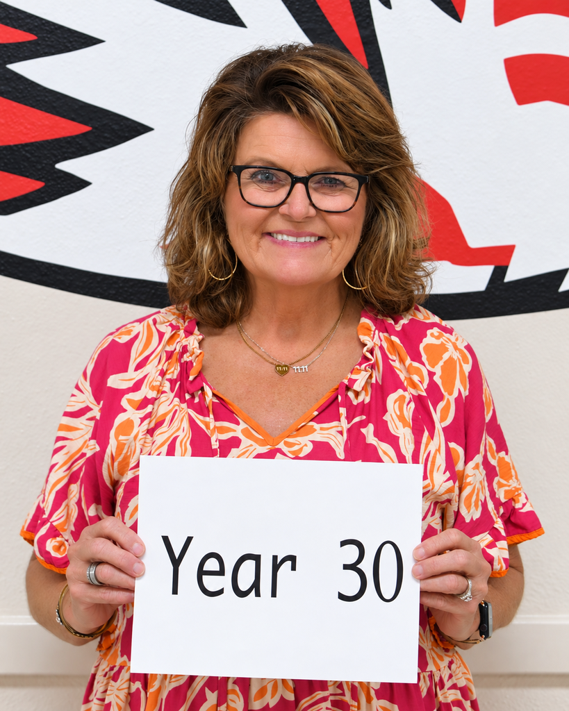 A woman in glasses stands in front of a wall mural, holding a sign that reads "Year 30."