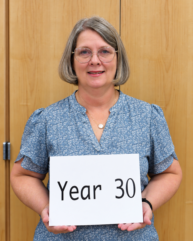Person with gray hair in a blue top holding a sign that says "Year 30". Background is wooden.