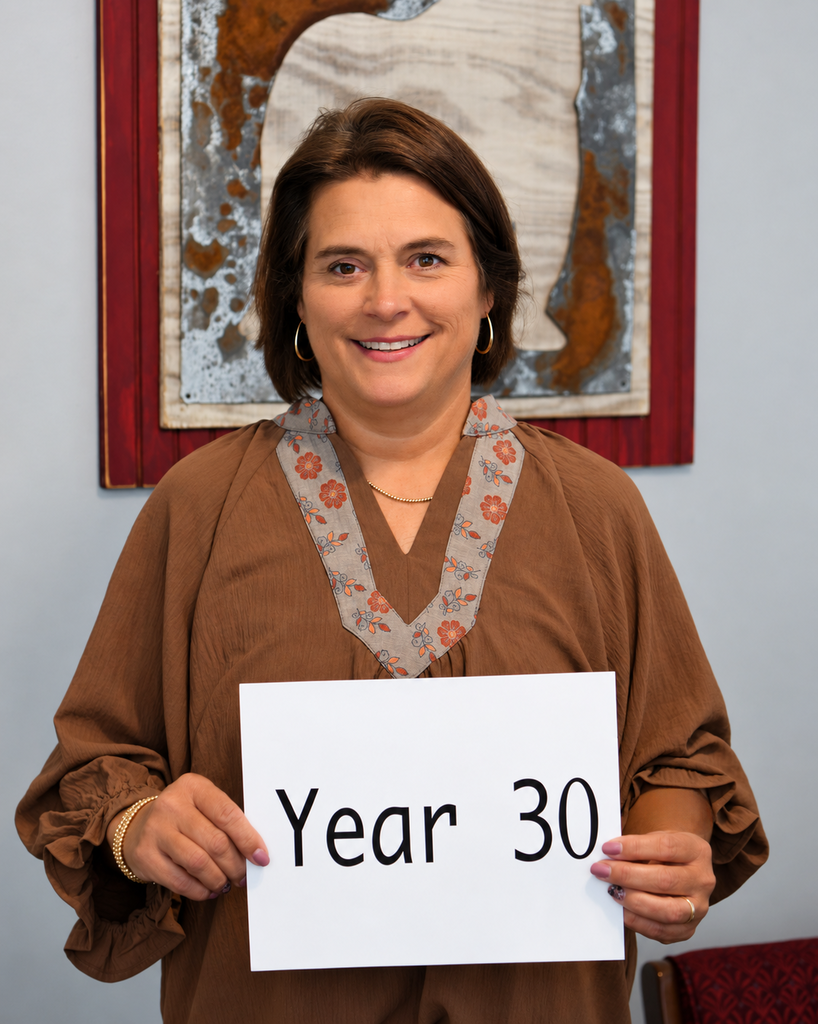 A smiling woman in a brown shirt holds a sign that reads "Year 30" in front of a red-framed wall art.