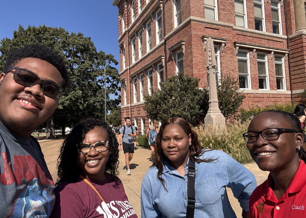 Four individuals standing in front of a red brick building, smiling. One wears glasses, another a blue shirt, and others wear maroon and red.