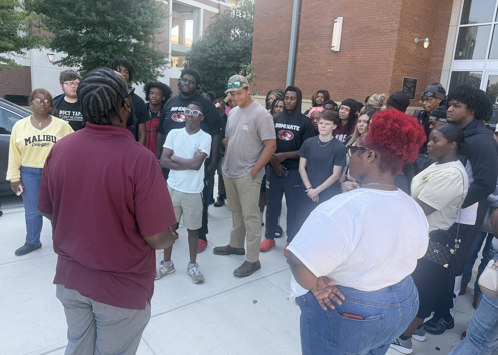 A group of young people gathers outside a building with brick walls. One person wears red hair, and another wears a cap.