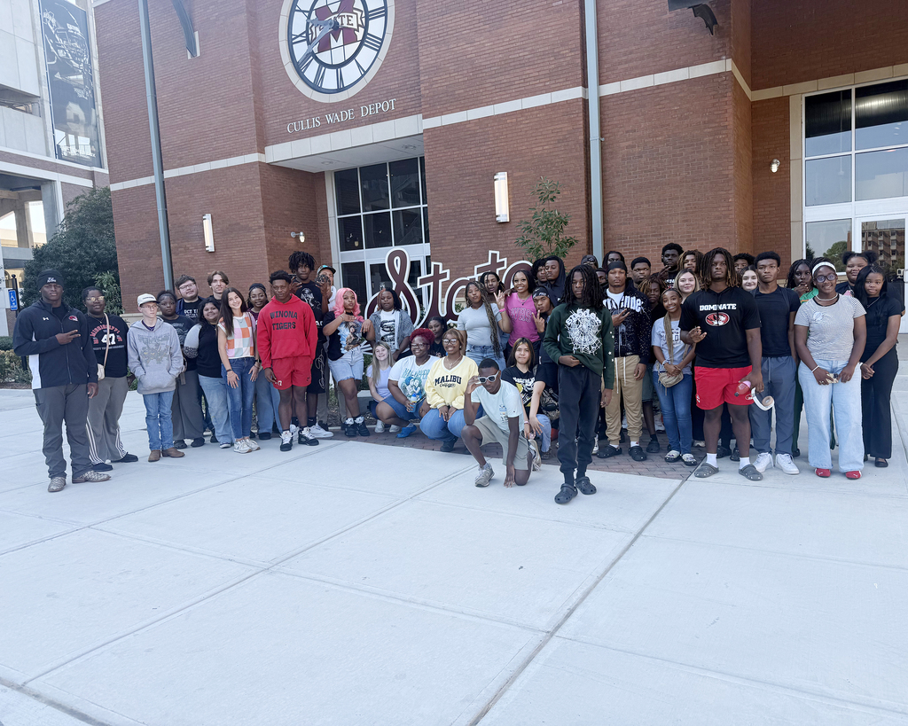 A group of individuals poses in front of a brick building with a clock on its facade.