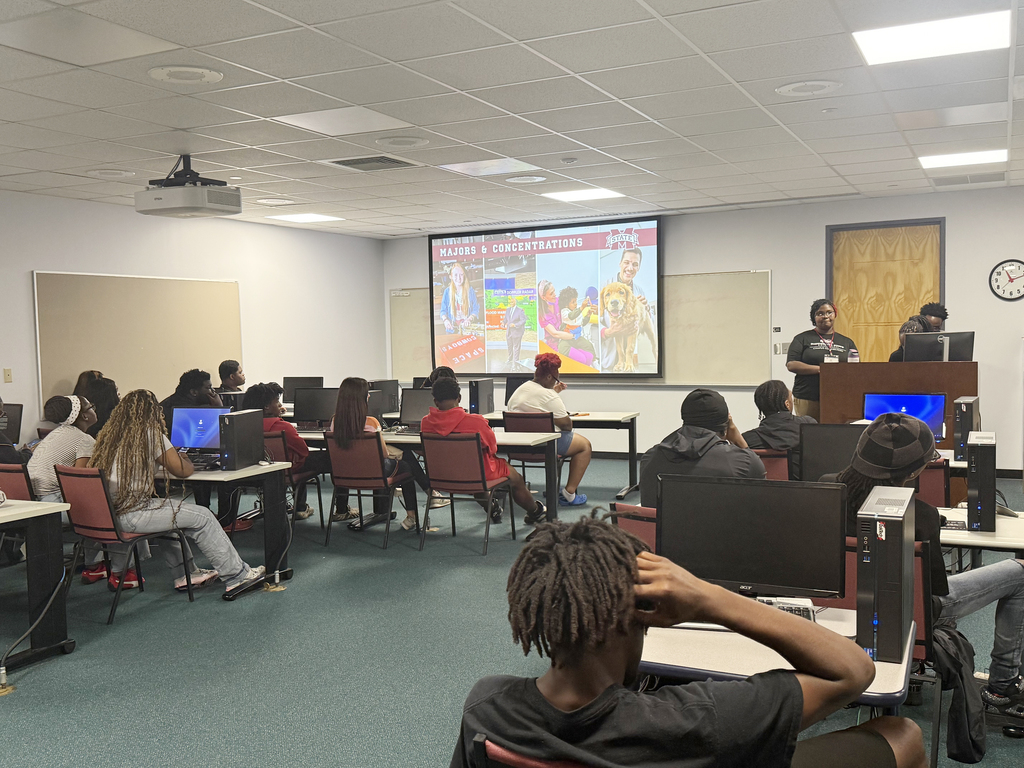 A classroom with students seated at desks, some wearing headphones, listening to a presentation on a projector screen.