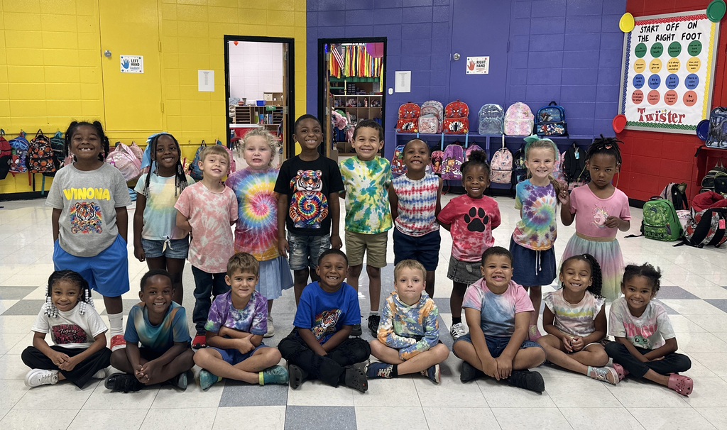 A group of children, wearing various T-shirts, gather for a classroom photo with colorful walls and backpacks in the background.