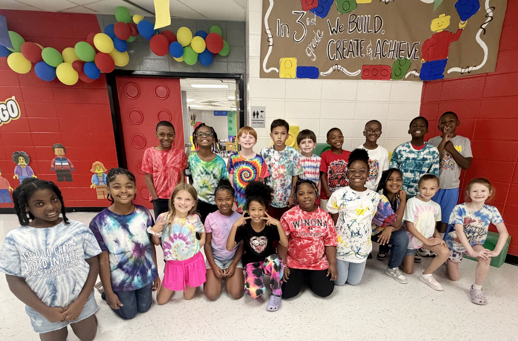 Children wearing tie-dye shirts and colorful clothes pose for a group photo in a classroom.