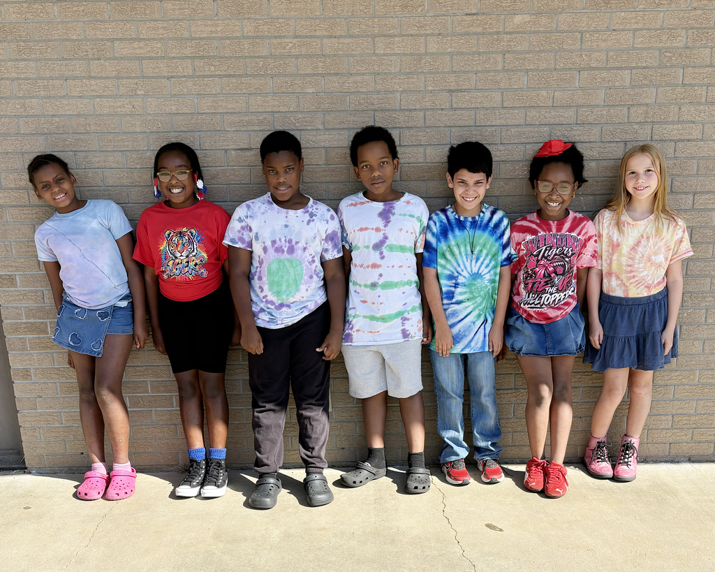 Seven children wearing colorful shirts stand against a brick wall, each with a unique shirt design, and different colored shoes.