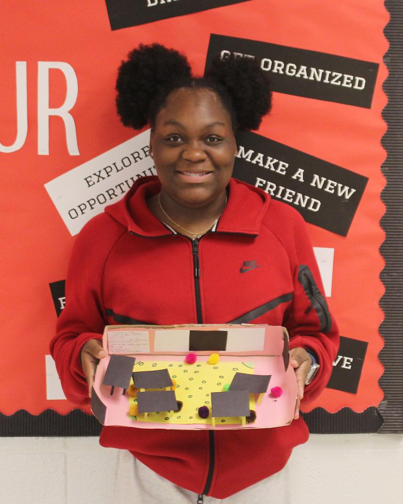 Woman with dark curly hair holds a pink box with yellow and black shapes. Behind her, red and black posters with white text.