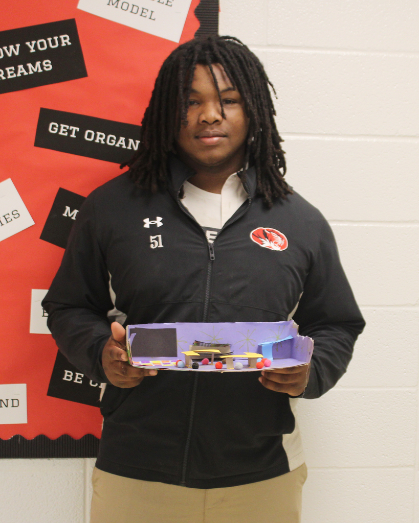 A person stands against a motivational banner, holding a small, purple model of a car.