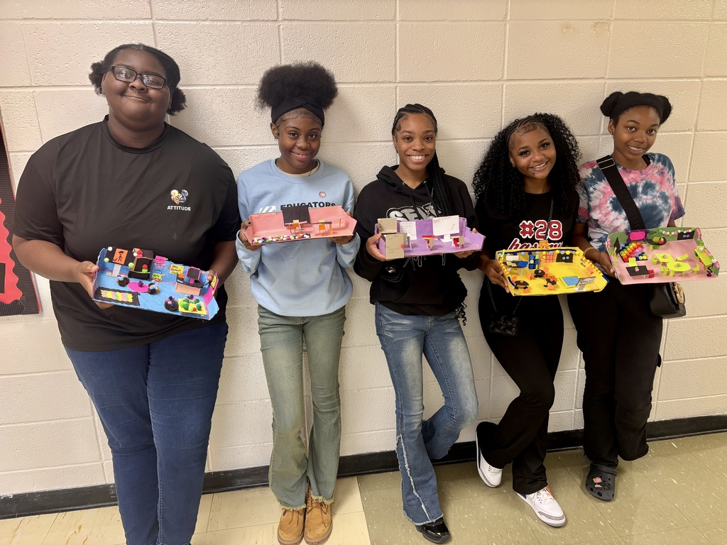 Five young women pose together, holding rectangular boxes with miniature structures inside. They stand against a white background.