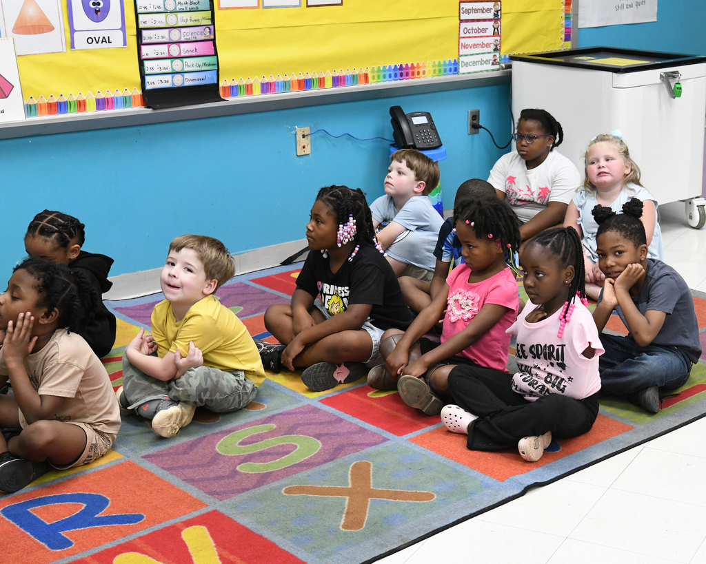 Group of children, ages 5 to 6, sitting on a colorful mat in a classroom with blue walls.