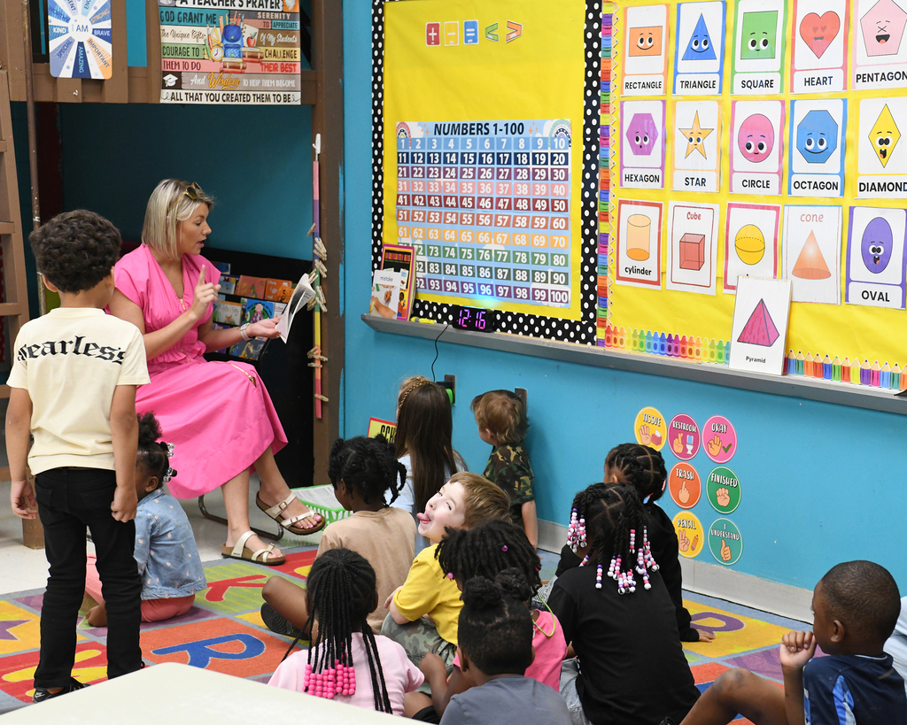 Woman teaching a classroom of young children; colorful bulletin board behind her.
