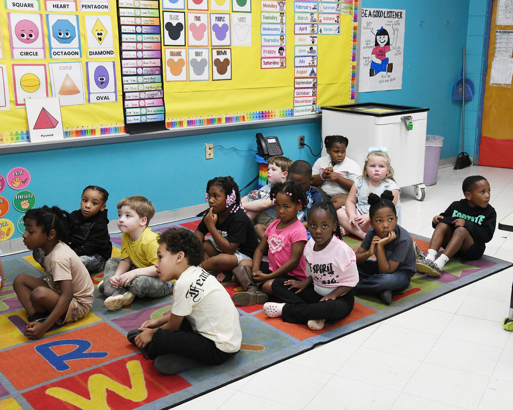 Group of young children sitting on carpet in a classroom. Behind them is a bulletin board.