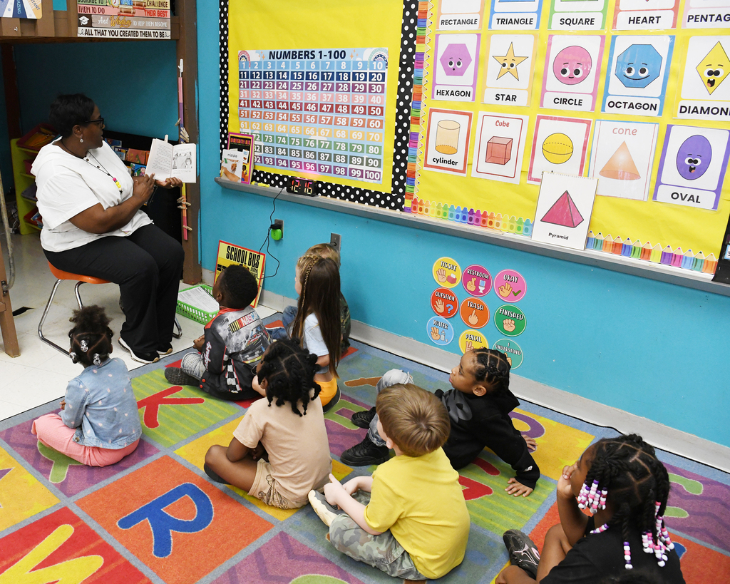 A classroom with a teacher reading to a group of children sitting on a rug. A colorful chart is on the wall.
