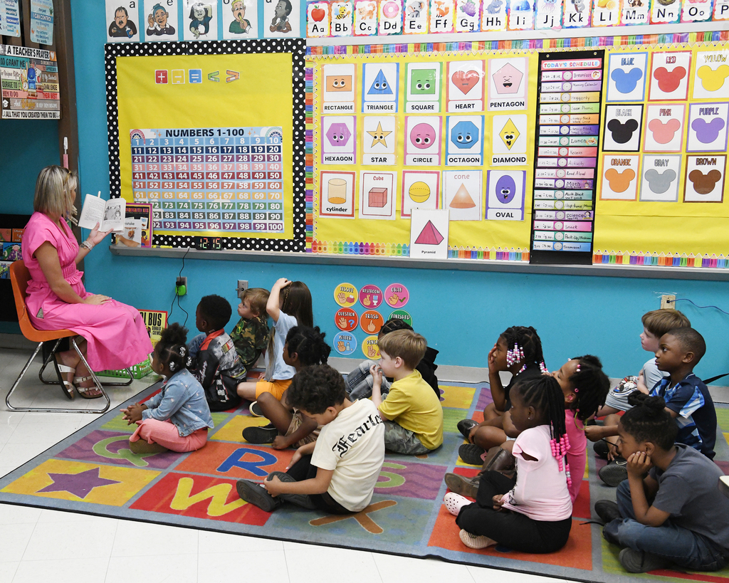 Woman reading a book to a group of children sitting on a colorful carpet.