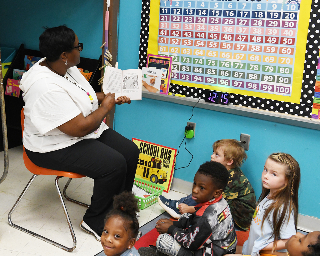 Woman reading to children in a classroom. Wall with number chart. Children sit on floor, listening.