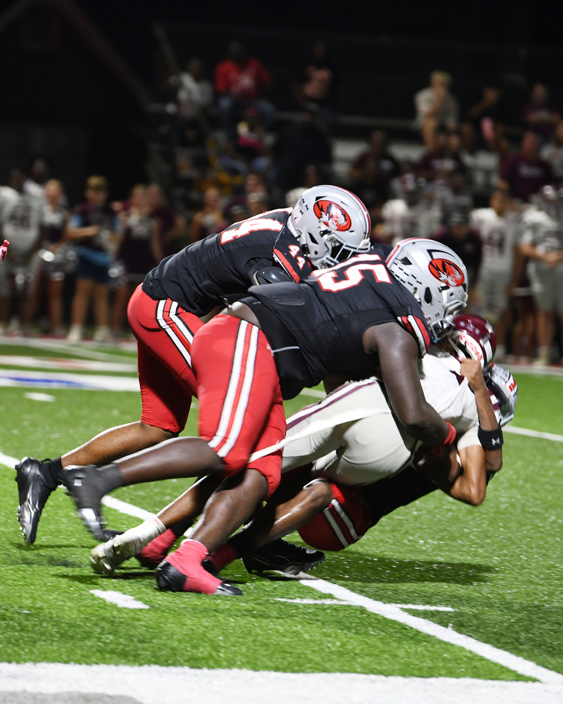 Players in jerseys and helmets engage in a football tackle on a field. Audience seated in stands watches.