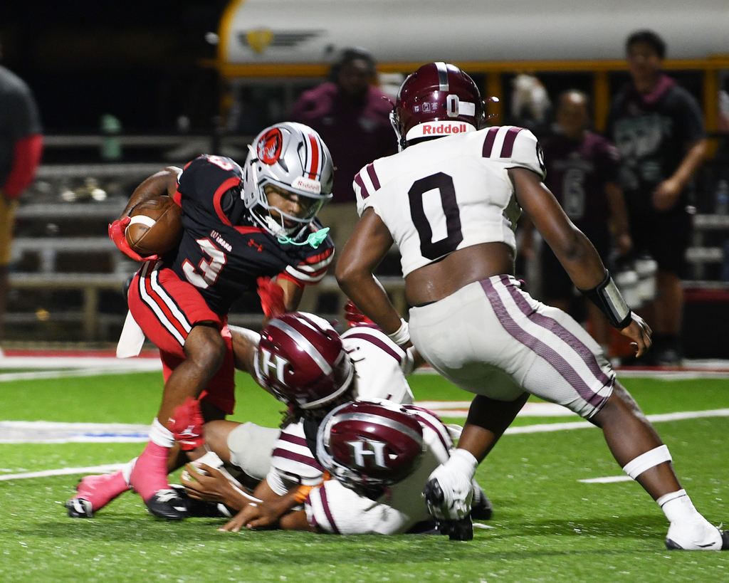 Players engage in a tackle during a football game, one player in red and white, another in white, and a third in maroon.