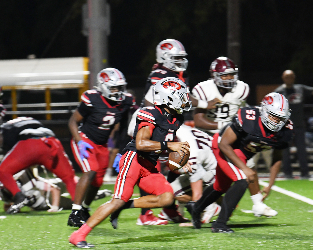 A group of football players on a field. Two teams in red and black jerseys. One player runs with the ball.