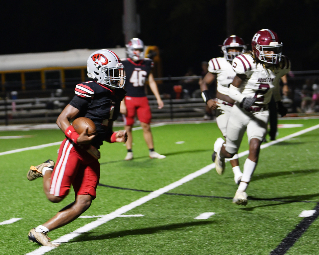 Players in football uniforms on a field. One running with a ball, others chasing. White lines and bleachers in the background.