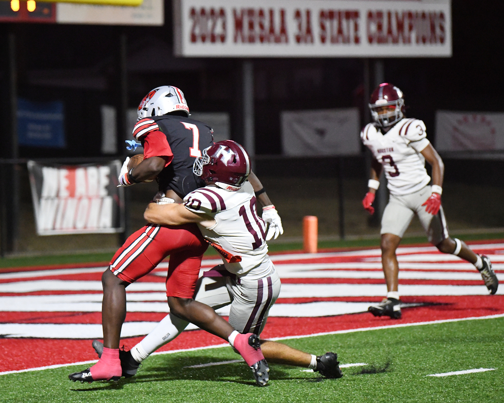 Two players wrestle on a field, one in red and the other in white. A player in maroon runs in the background. Advertisements and banners in the background.