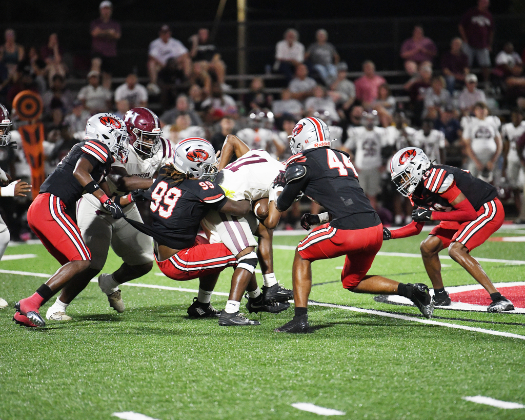 Football players in black and red uniforms tackle each other on the field. Audience seated on stadium benches in the background.