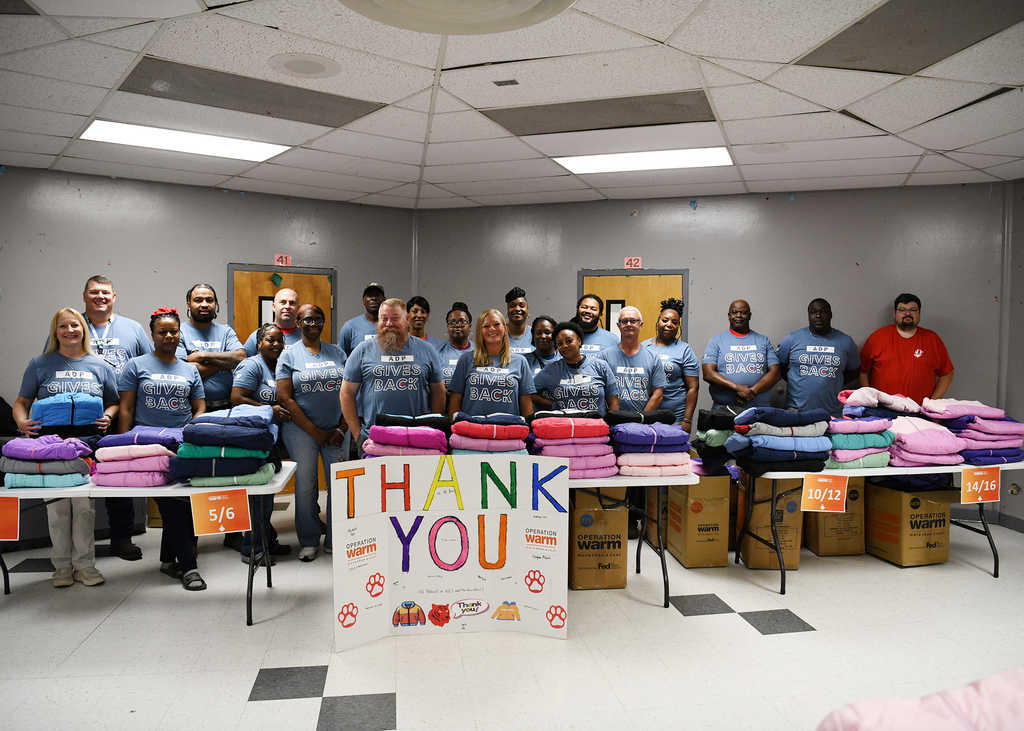 A group of people in matching blue shirts stands behind tables of blankets. A white sign reads "Thank You".
