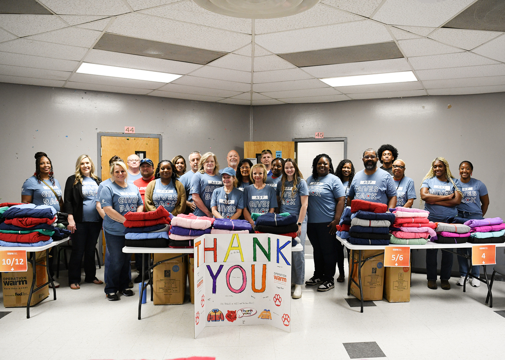 A group of people in matching shirts stand behind tables stacked with folded clothes, near a sign that reads "THANK YOU."