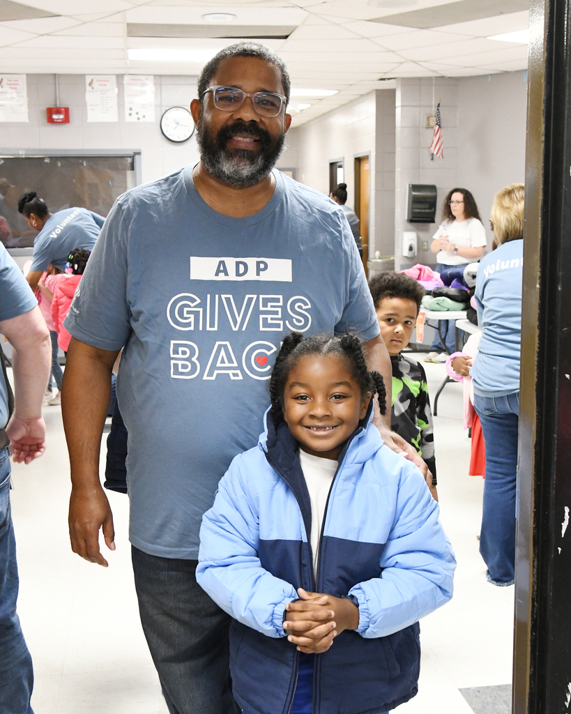 A man with a beard and glasses stands with a smiling child in a blue coat. They are in a hallway.
