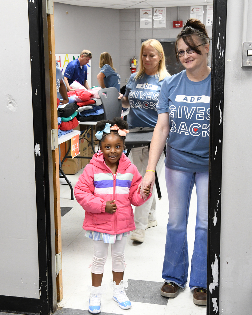 A woman helps a young girl walk through an open door. Behind them, other people sort through clothing.