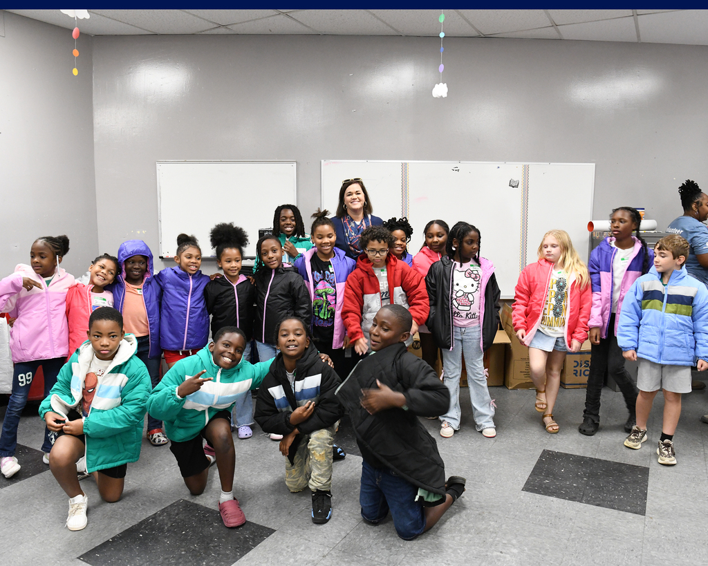 Group of children and adults standing and kneeling in a room with whiteboards on the wall.