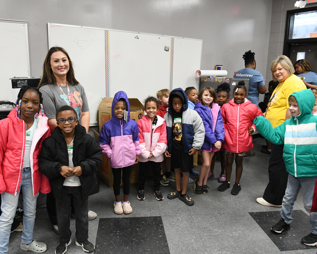 A group of children and two women stand in a classroom. The children wear various jackets and sneakers.