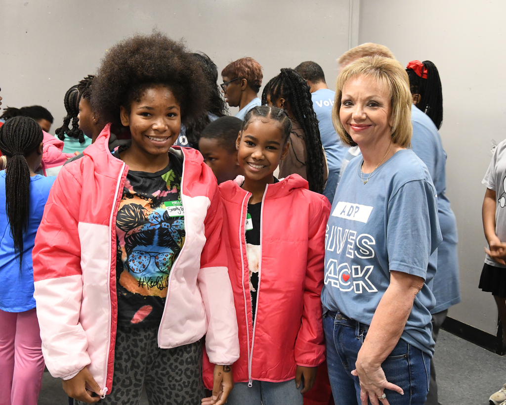 Three women in jackets stand next to each other with children. The woman on the right smiles with her hands on her hips.