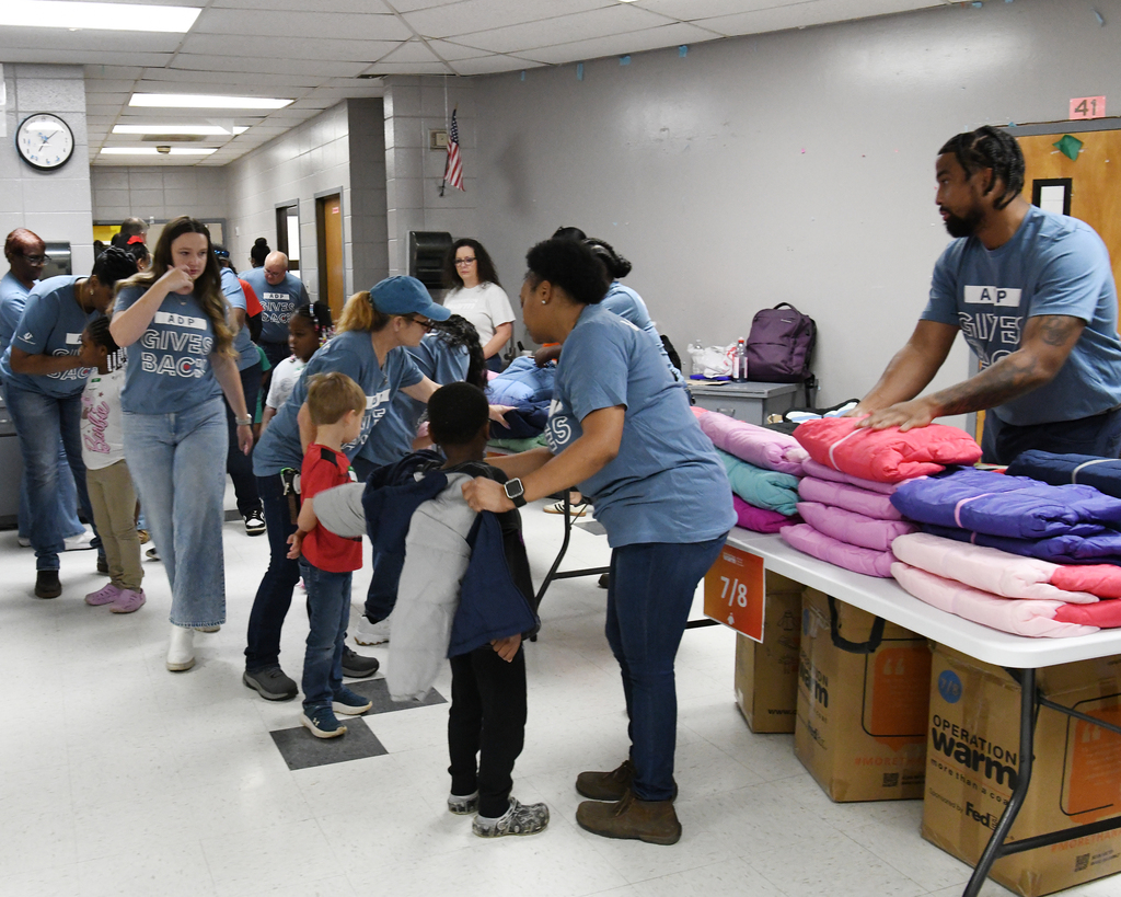 A group of people in blue shirts are in a hallway, helping children with clothing at a table.