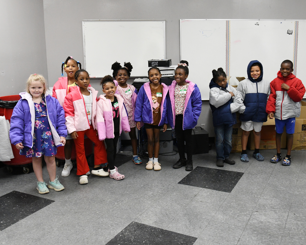 A group of children, some wearing jackets, in a room with white boards and a trash bin.