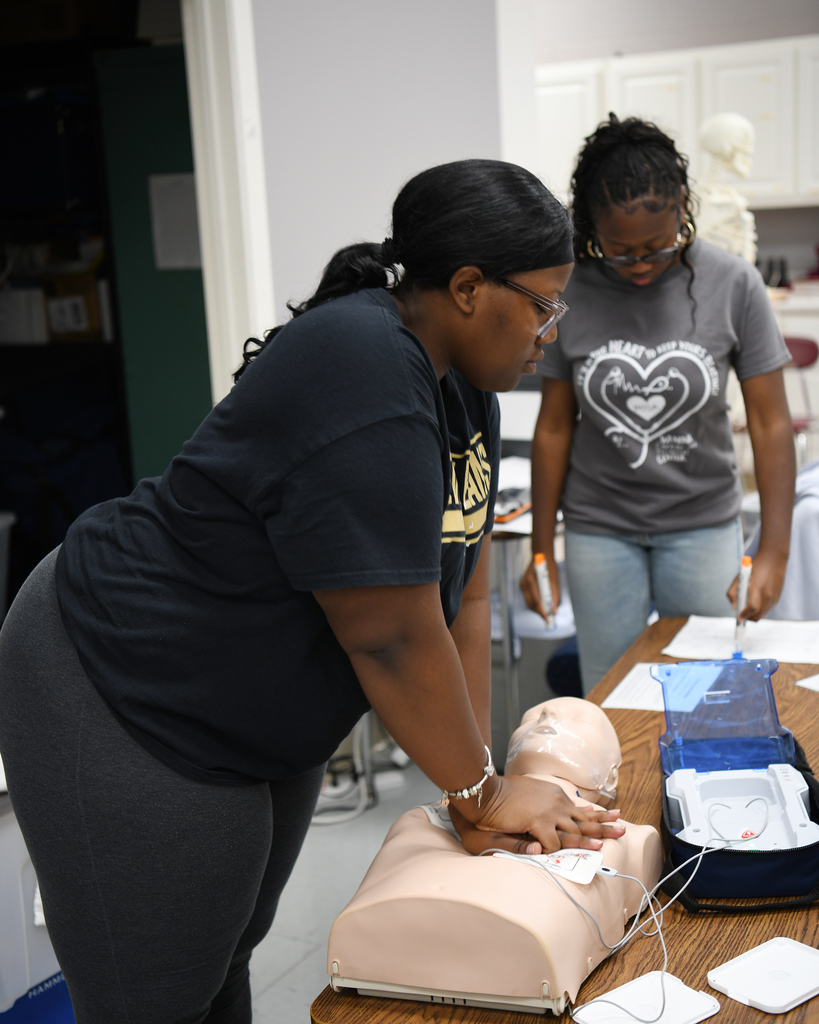 Woman in black shirt uses manikin to practice CPR as another person watches. Background includes cabinets and a mannequin.