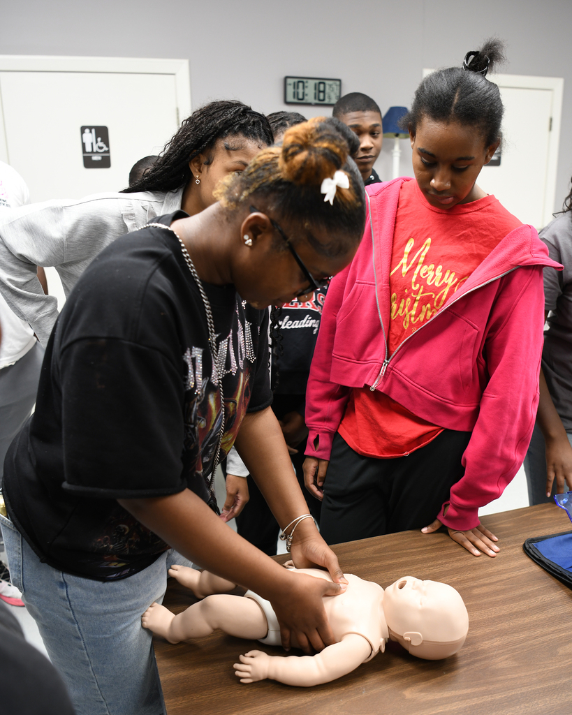 People gathered around a table, one person wearing a black shirt bends over a baby doll lying on the table.