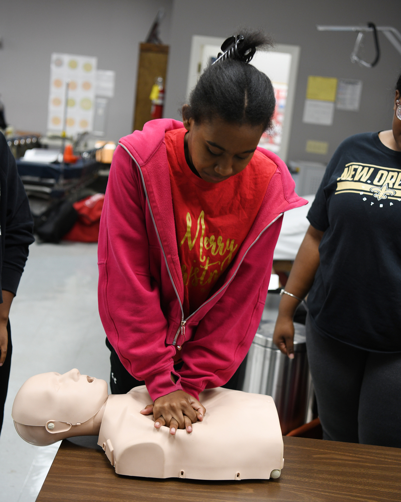 A woman in a pink hoodie demonstrates CPR on a dummy while a person in black observes.