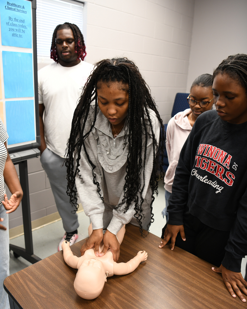 Students in a classroom setting with a woman examining a CPR training mannequin on a table.