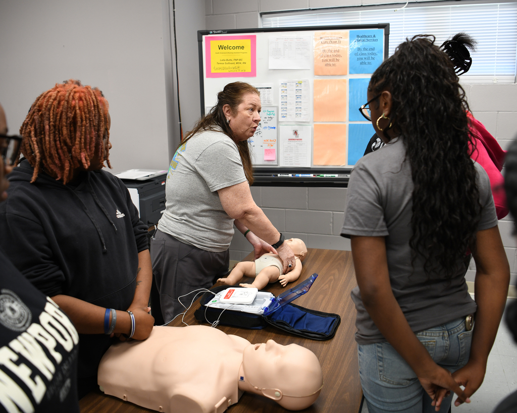 A group of people in a room with a board on the wall. A woman demonstrates CPR on a mannequin.