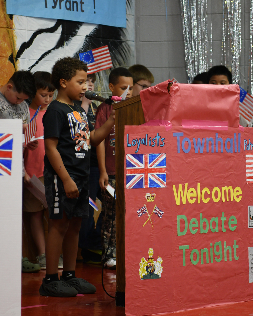 Group of children in a classroom setting, one standing at a podium holding a microphone. Nearby, a banner displays "Townhall Welcome Debate Tonight" and flags of the UK and US.