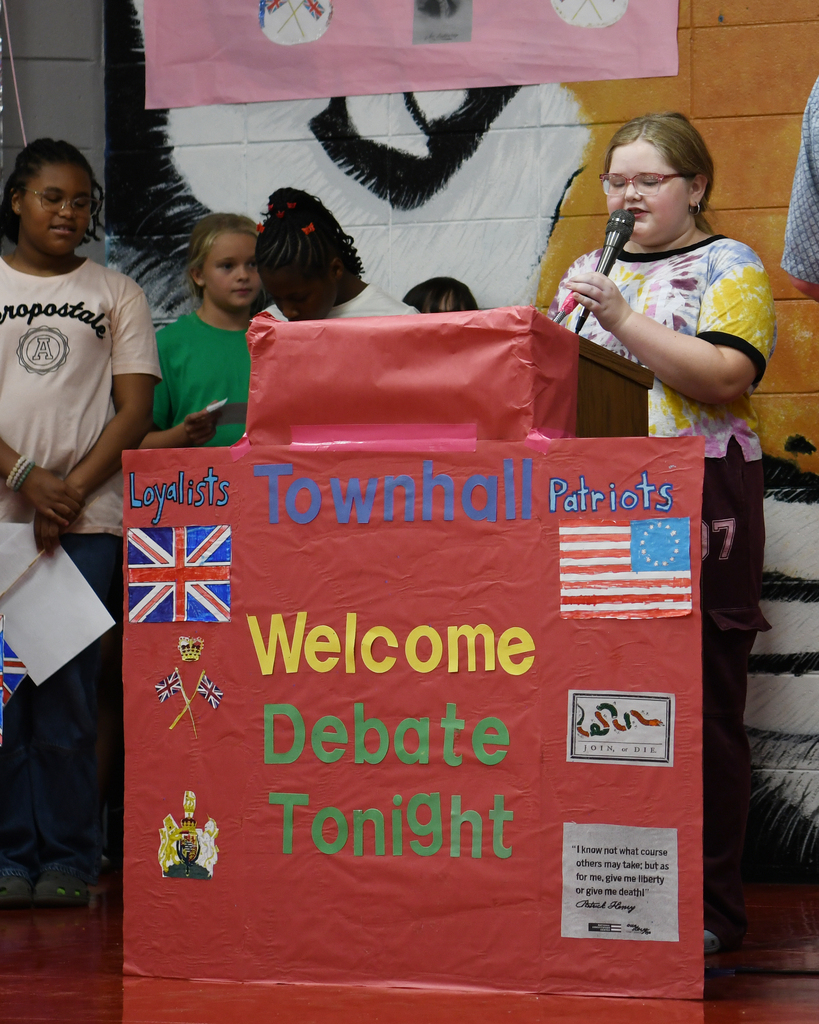 A young girl stands at a podium, addressing a crowd. A sign behind her reads "Townhall Patriots Welcome Debate Tonight."