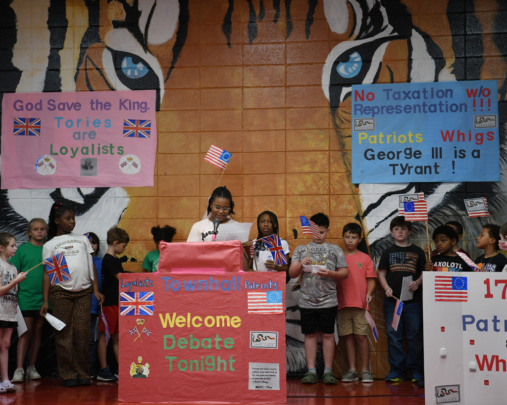 Children holding flags stand in front of a wall with a tiger and political posters.