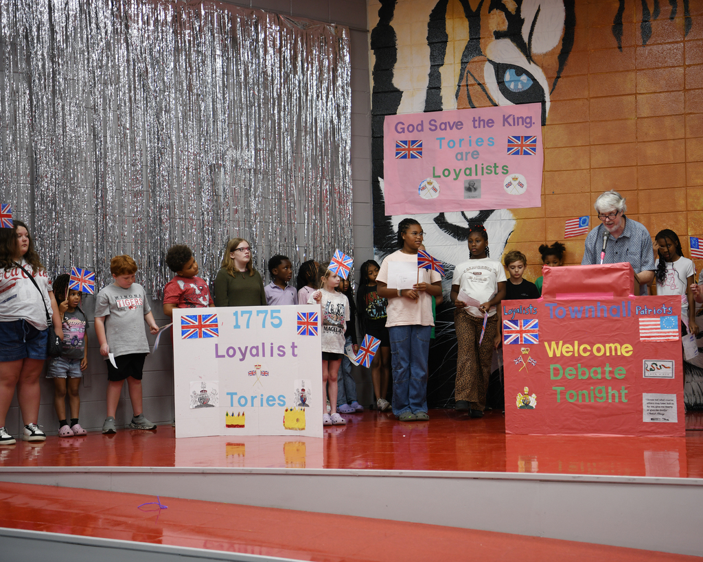 Kids stand on stage with signs, one holding a sign with "1775 Loyalist Tories". Behind them, a backdrop with a tiger and a sign reading "God Save the King Tories are Loyalists".
