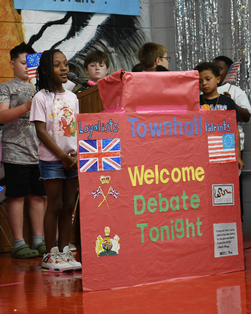 A group of children stand by a red sign reading "Townhall Welcome Debate Tonight" with flags of the UK and USA.