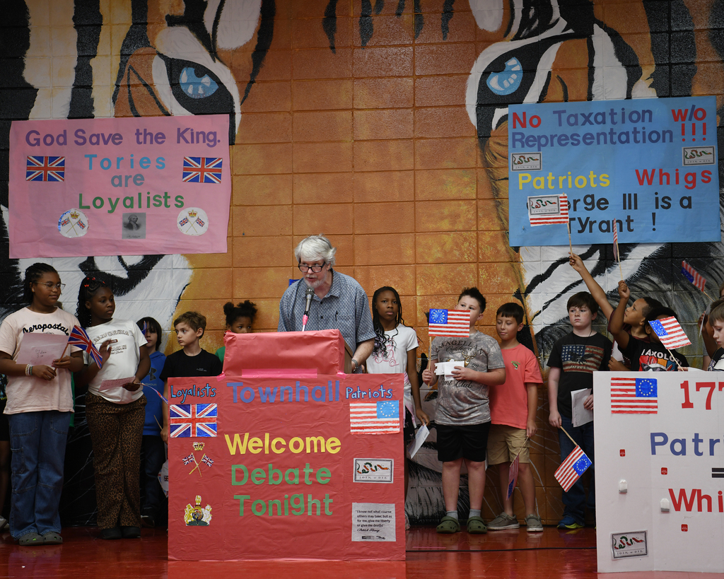 Children in a school gym debate. A man speaks at a podium. Two banners behind them read "God Save the King" and "No Taxation Without Representation".