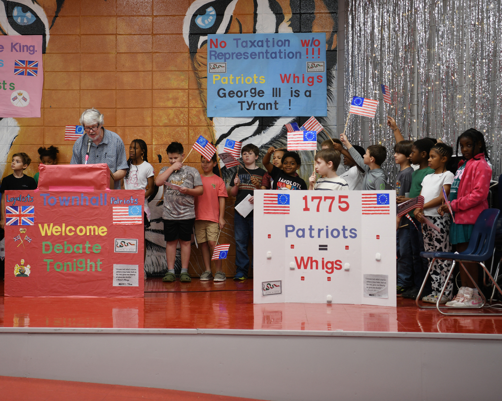 Children holding flags stand on a stage with signs and banners; a man at a podium speaks.