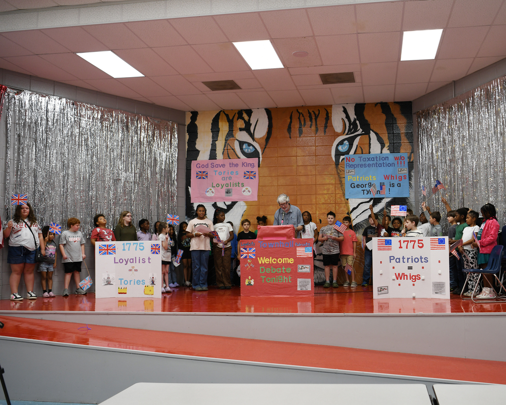 Group of children on a stage with signs. Two banners show flags. Tiger graphic on wall behind.