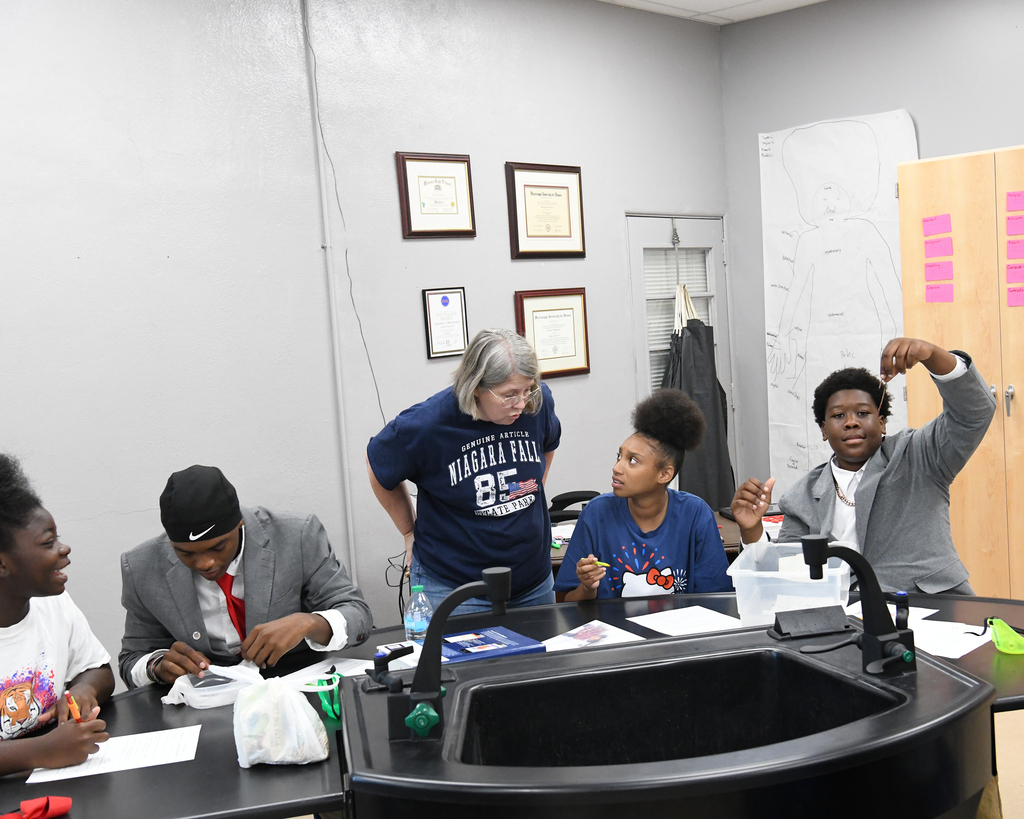 Several students in a lab setting, two are raising hands, and one is standing behind a table.