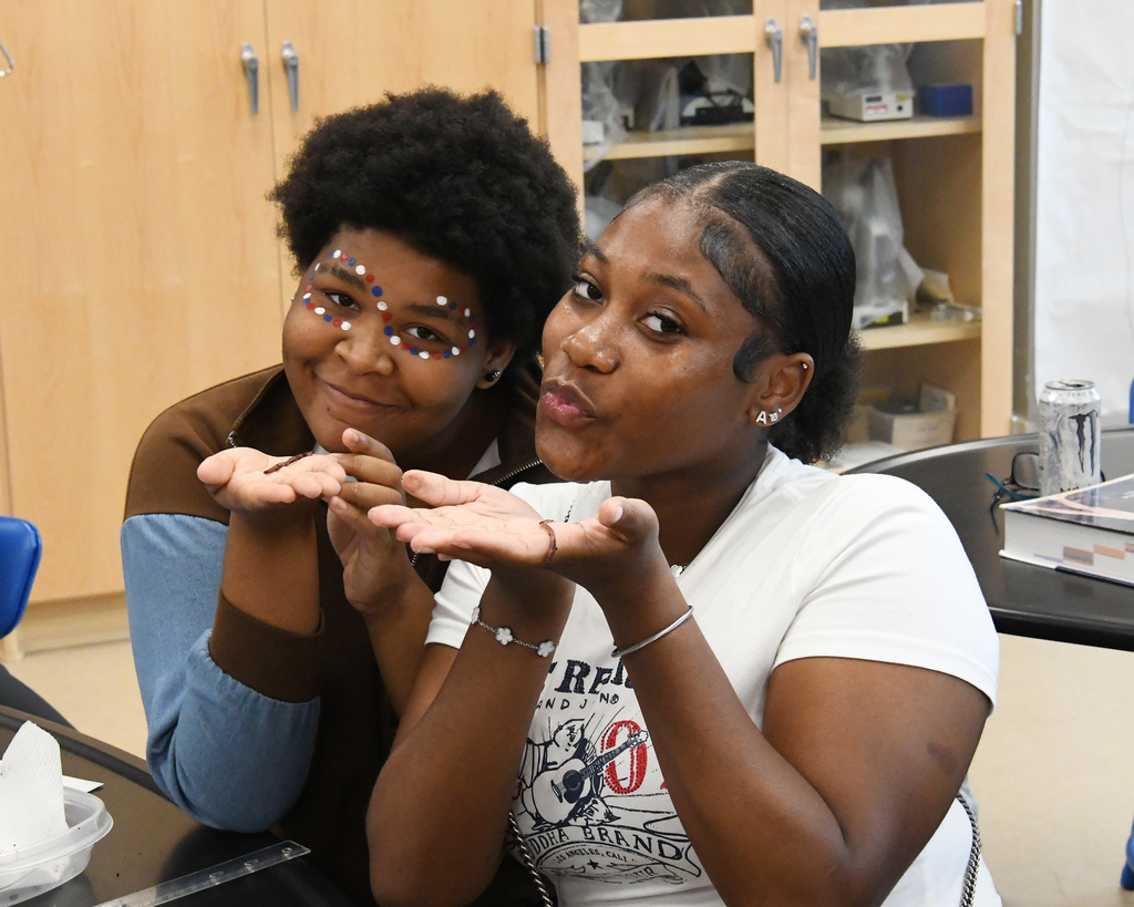 Two women seated, one with face paint, gesturing while looking at something in a lab room.