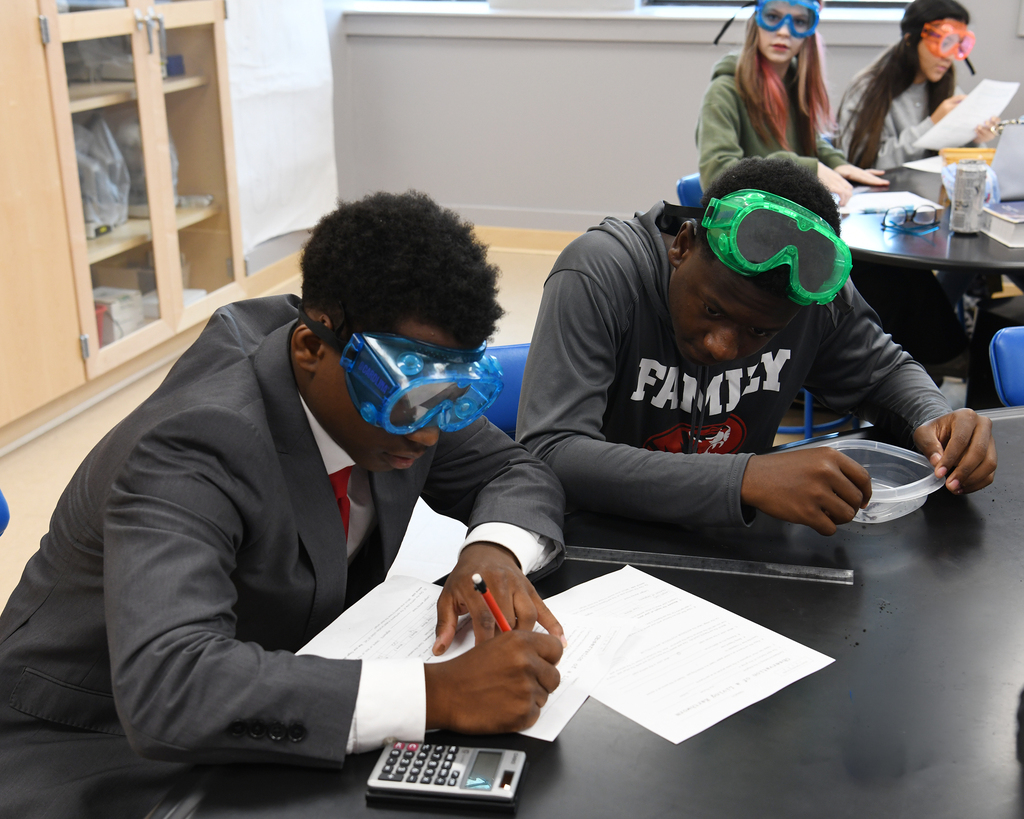 Two students wearing goggles, one writing on paper, another with a calculator, seated at a desk.